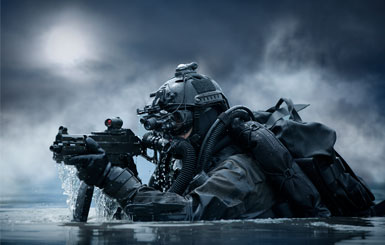 A combat diver with night vision googles and a submachine gun at the ready emerges from the sea.